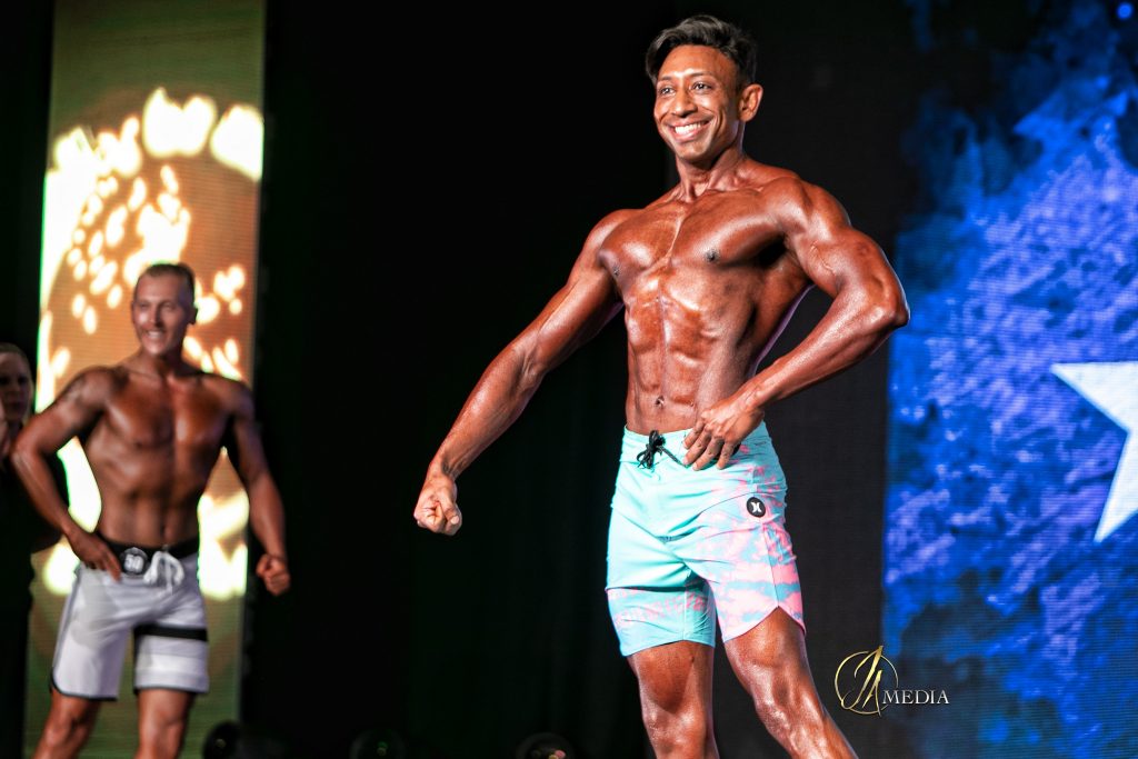 &ldquo;Dr. Shiv Goel confidently posing on stage at an IFBB men&rsquo;s physique competition, showcasing the results of disciplined training and holistic wellness.